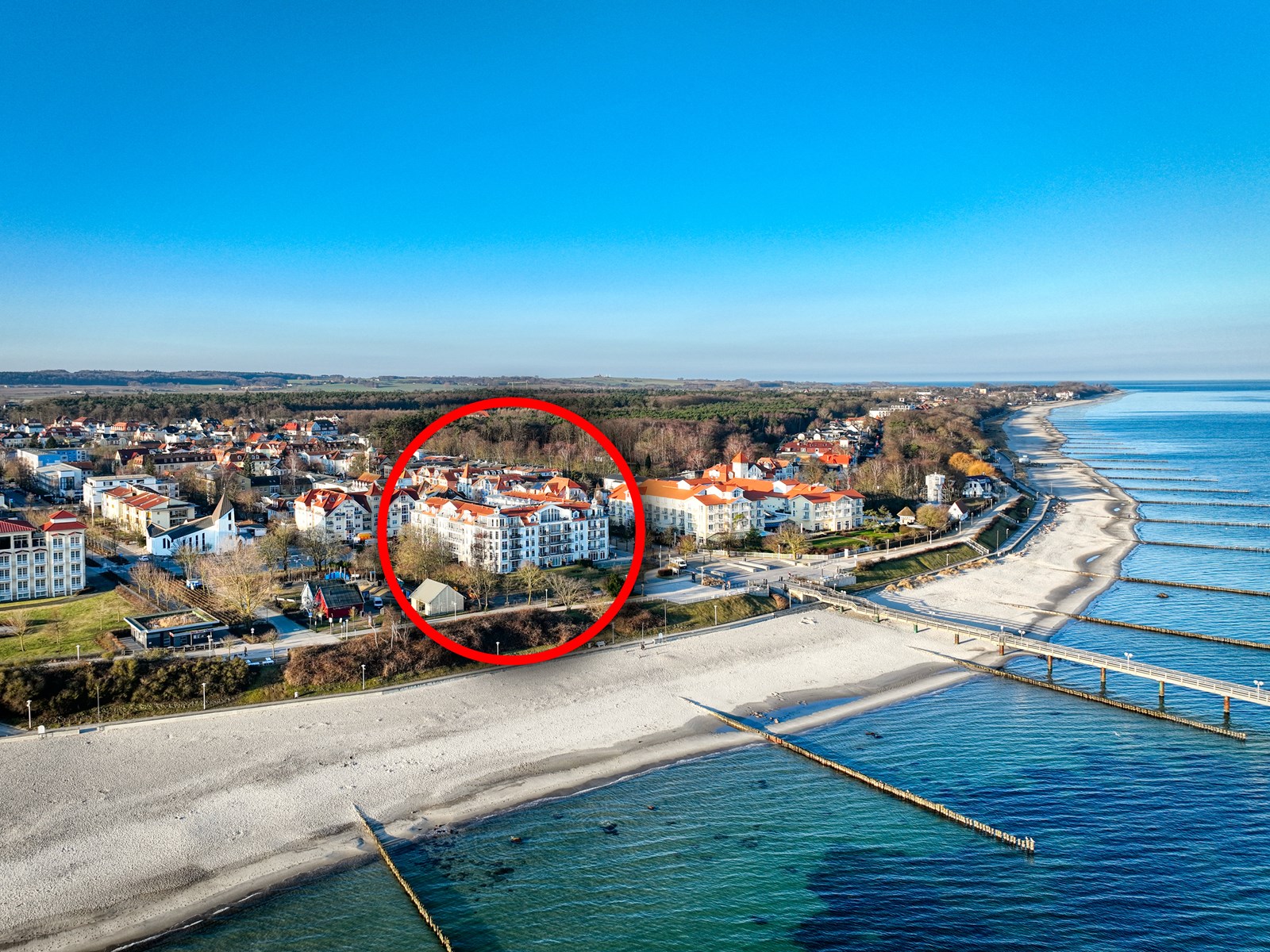 Aerial view of a white apartment building with orange roofs, circled in red, near a sandy beach and blue ocean.