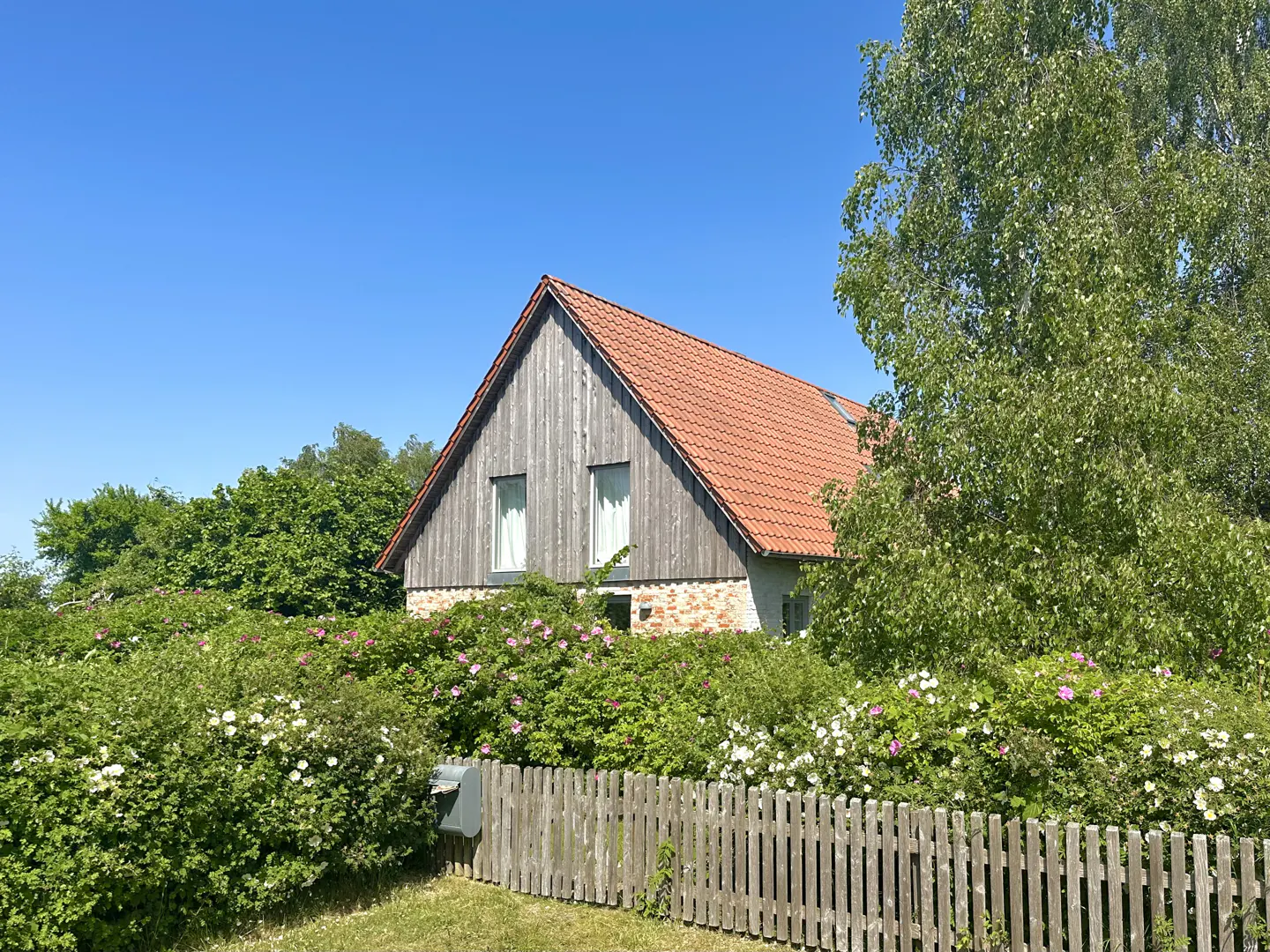 Exterior view of a house with a red tile roof, gray wood siding, surrounded by green bushes and a wooden fence.