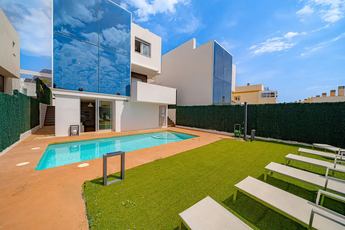 Modern white house with a pool, green lawn, and lounge chairs under a blue sky. The house has a blue glass facade.