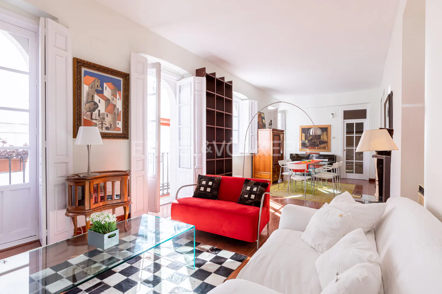 Bright living room with white sofa, red loveseat, and glass table. Black and white checkered rug on wood floor. Open white doors lead to a balcony.