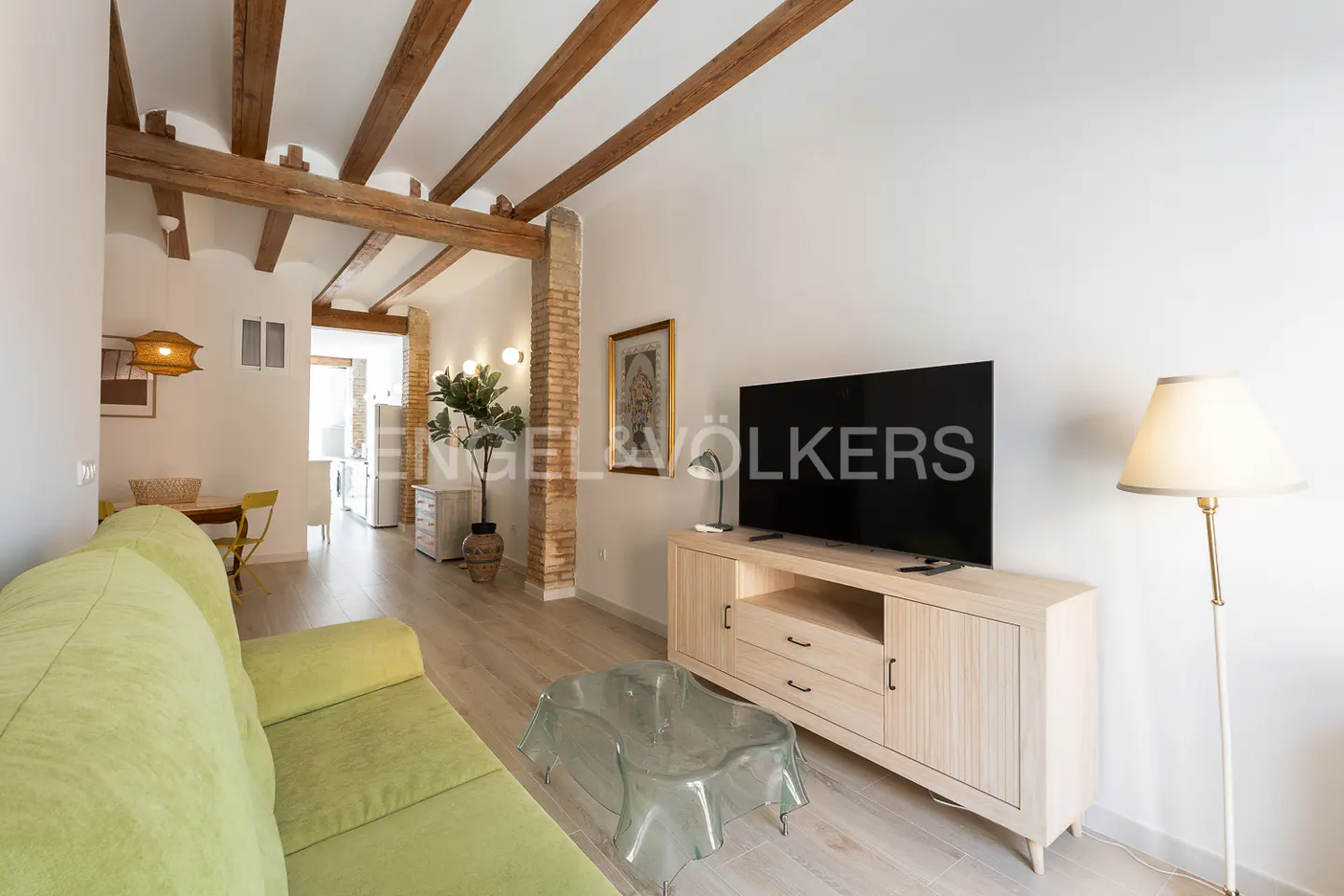 Living room with a green sofa, light wood floors, and exposed wood beams on the ceiling. A TV sits on a light wood cabinet.