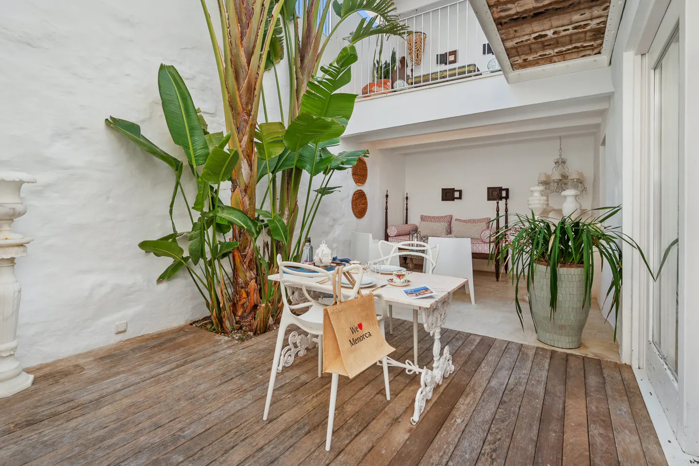 Indoor patio with white walls, wooden floors, and a white table set for tea. A "I love Menorca" bag hangs on a chair. Plants add a touch of nature.