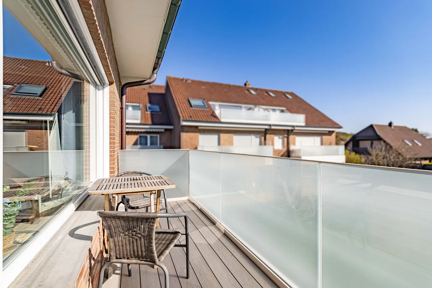 Balcony with a wooden table and chairs, frosted glass railing, and view of houses under a blue sky.