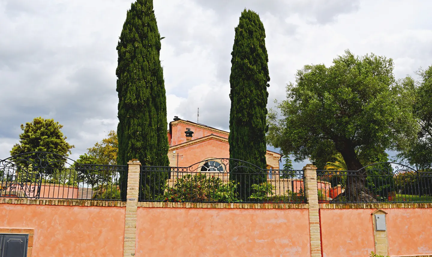A peach-colored villa behind a matching wall and black iron fence, framed by two tall cypress trees under a cloudy sky.