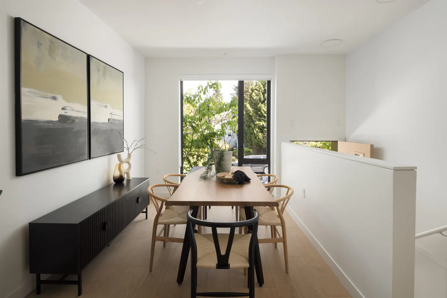 Bright dining room with a wood table, six chairs, and a black cabinet under two abstract paintings. A window overlooks green trees.