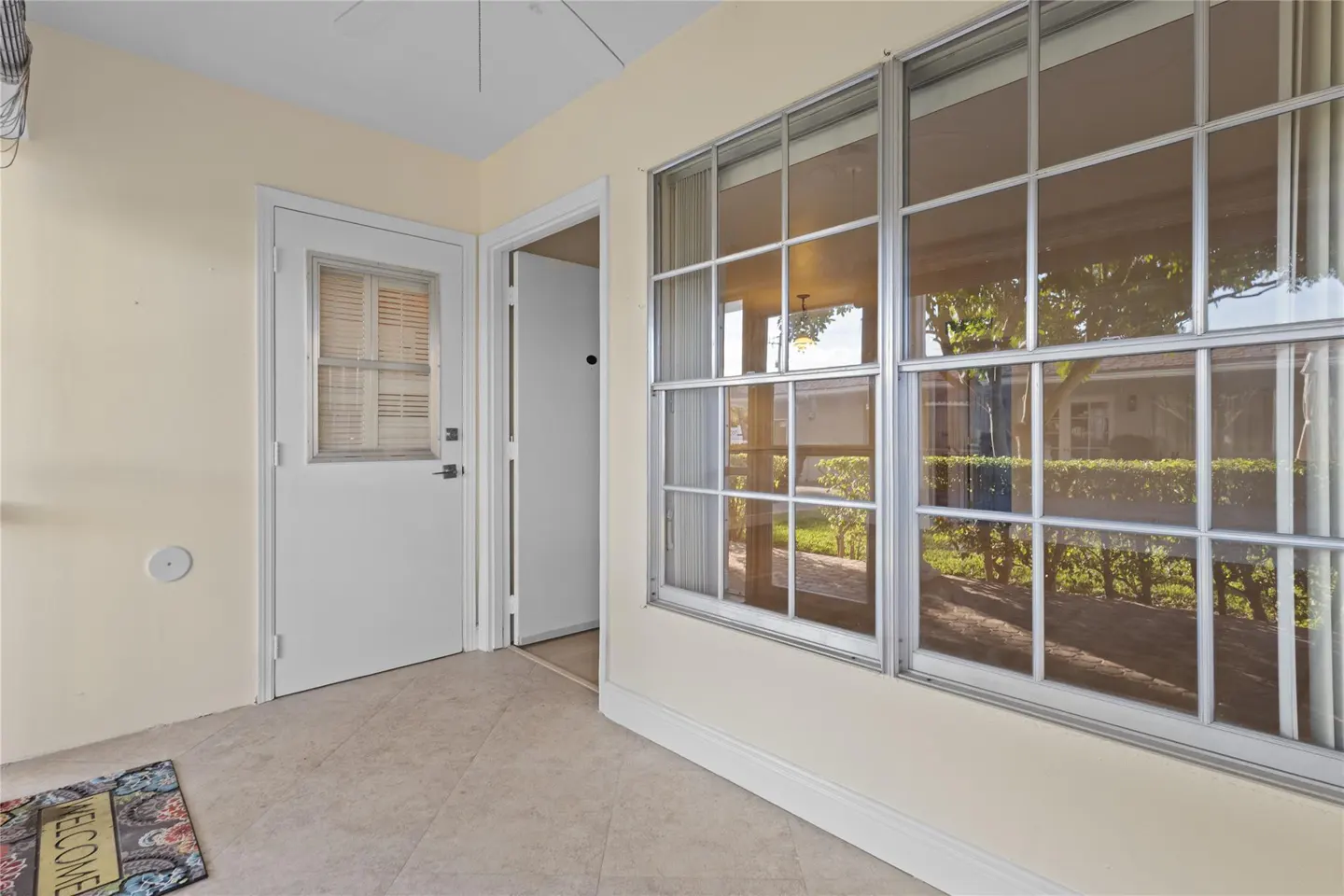 A sunroom with beige walls, a white door with blinds, and large windows showing a garden view. A welcome mat sits on the tiled floor.