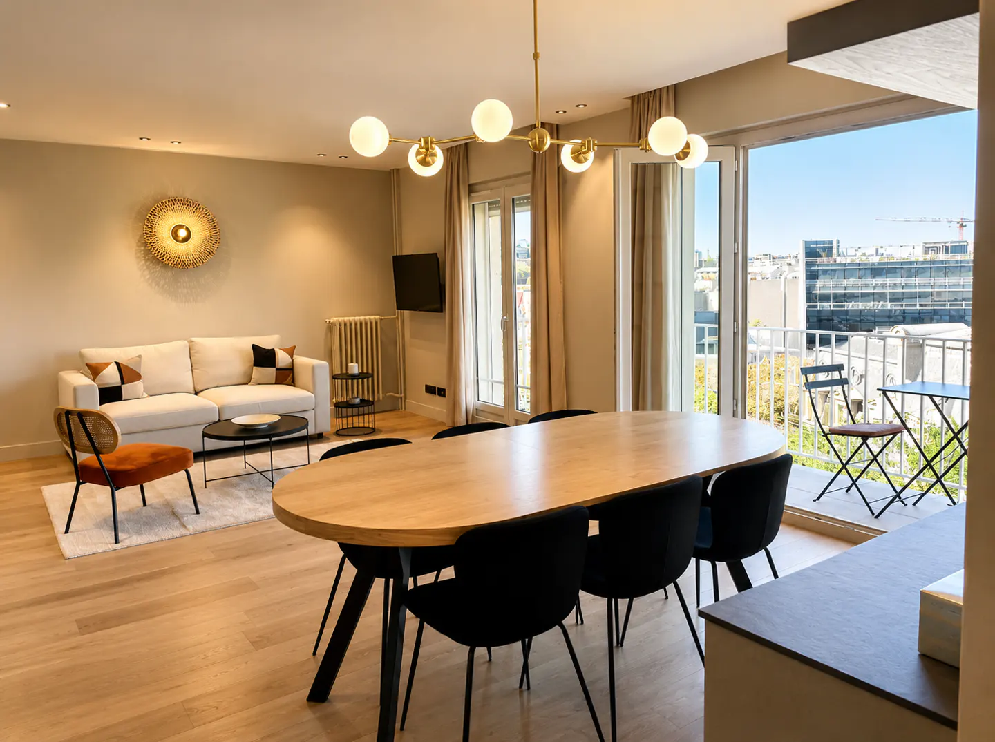 Bright living room with a wood table, black chairs, and a white sofa. Balcony doors open to a city view.