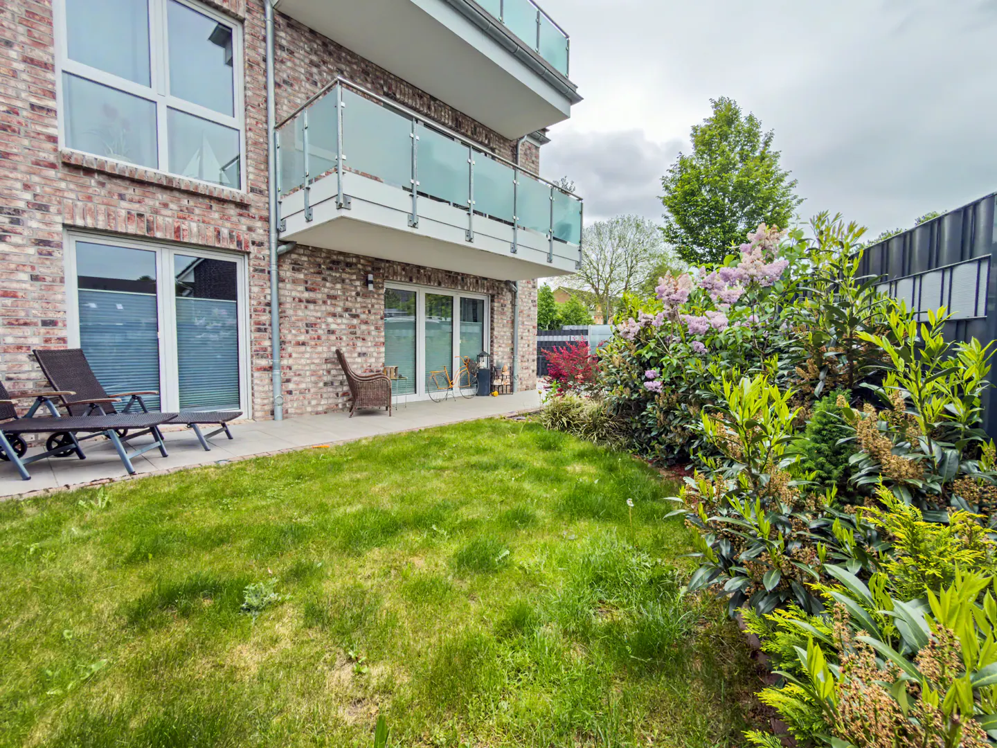 Modern brick apartment building with a balcony, patio furniture, and a lush green garden with flowering shrubs.