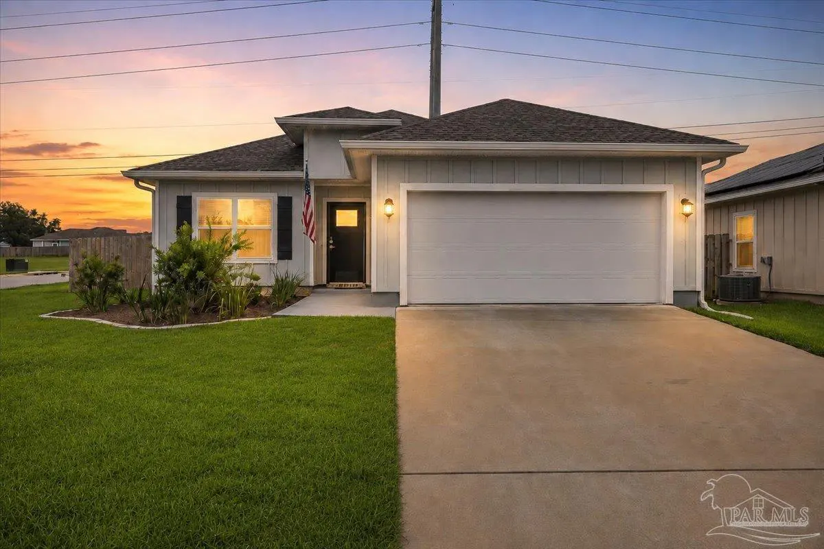 Single-story light gray house with a two-car garage, green lawn, and American flag at sunset.
