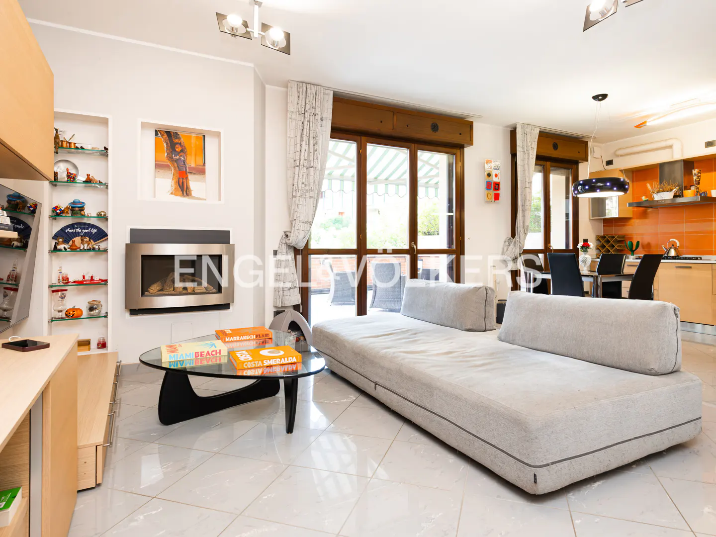 A bright living room with a gray sofa, glass coffee table, fireplace, and open kitchen in the background.