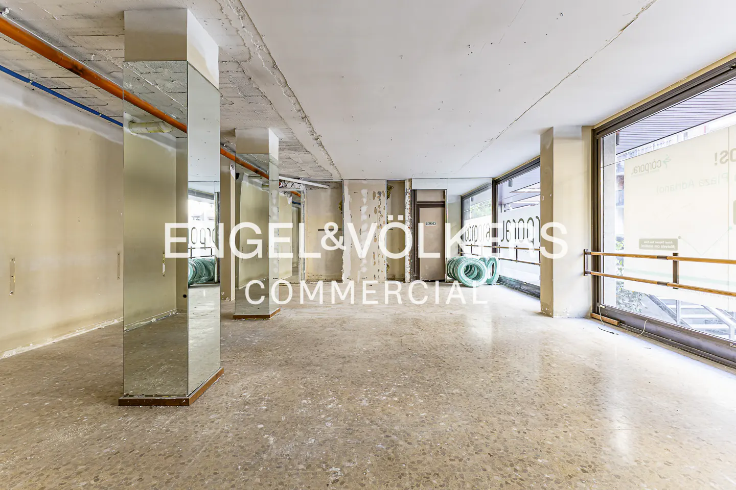Empty commercial space with mirrored pillars, large windows, and a speckled floor. "Engel & Völkers Commercial" logo overlay.