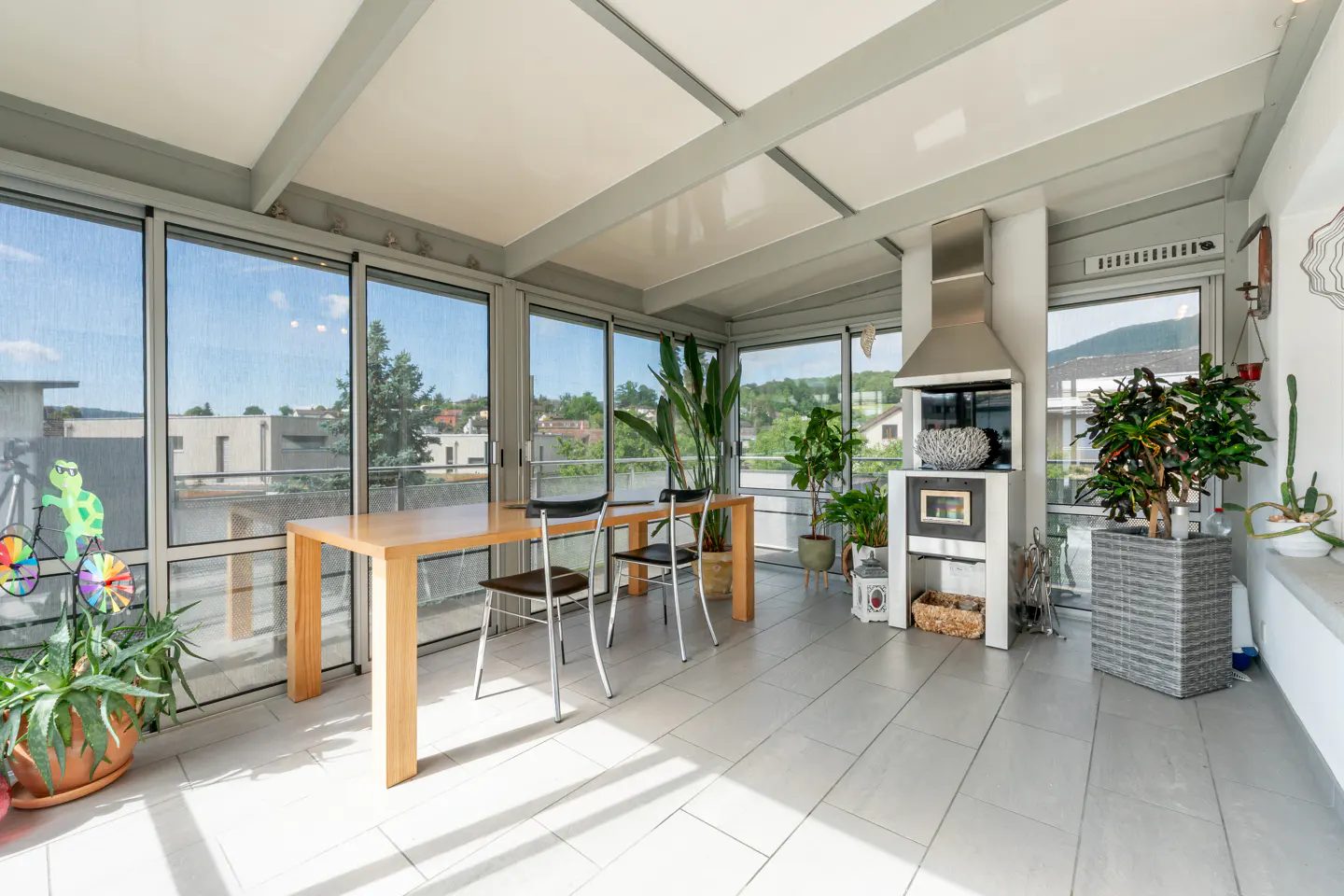 Sunroom with a wooden dining table, modern fireplace, tiled floor, large windows, and various potted plants.