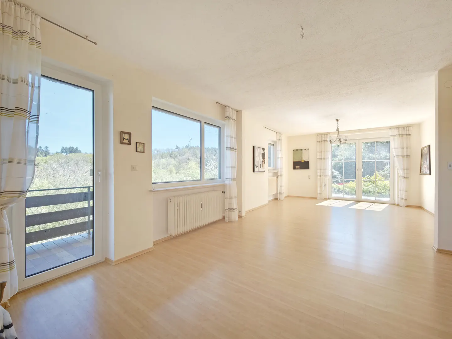 Bright, empty living room with light wood floors, white walls, and large windows with sheer curtains. Balcony and green landscape visible.