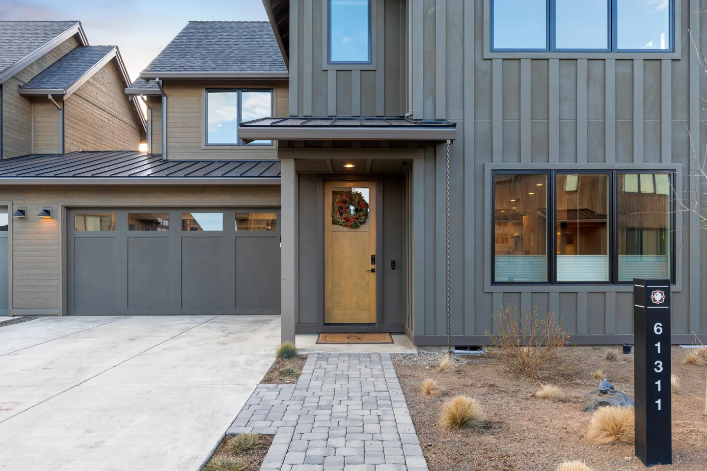 Exterior of a modern two-story house with gray siding, a wooden door with a wreath, and a gray garage door. A stone walkway leads to the entrance.