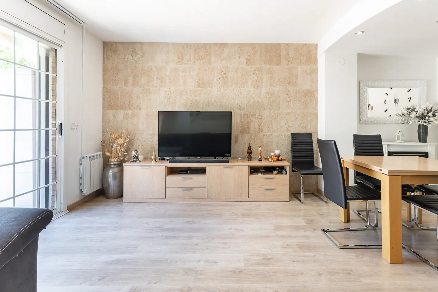 Living room with a TV on a light wood cabinet against a textured beige wall. A dining table with black chairs is visible. Sliding glass doors to the left.