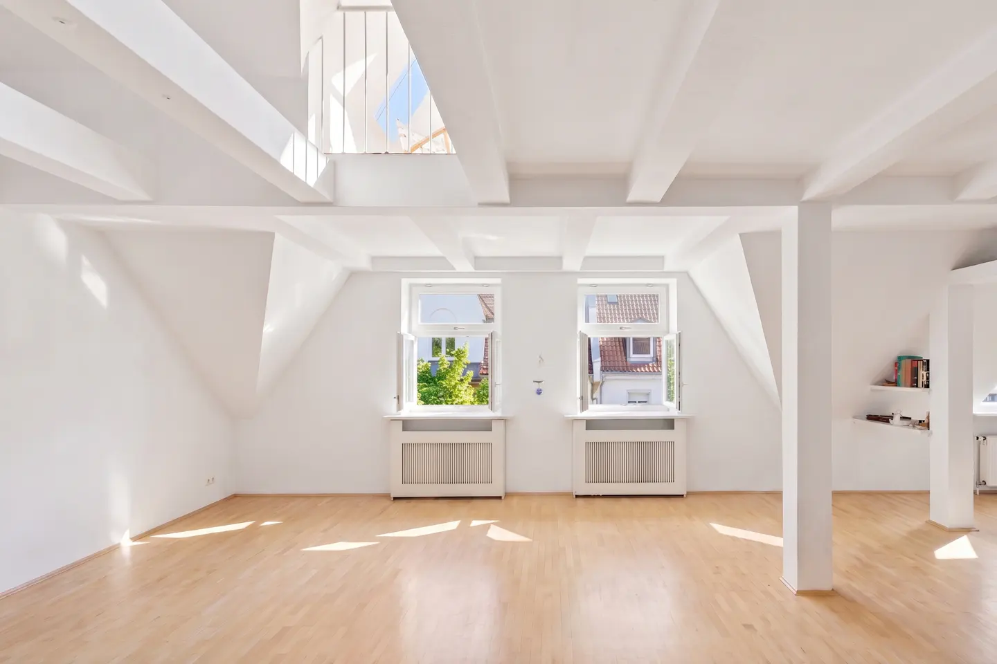 Bright, empty attic room with white walls, wooden floors, and exposed beams. Two open windows show trees and a building outside.