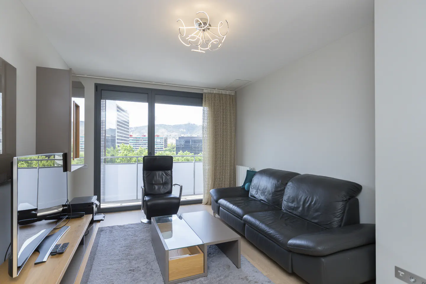 Living room with black leather sofa and chair, glass coffee table on gray rug. Balcony with city view. Modern light fixture.