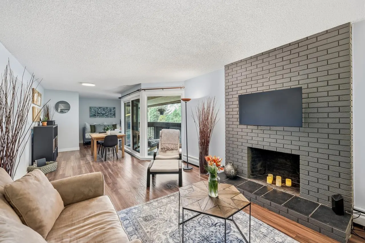 Living room with a gray brick fireplace, TV, and candles. A beige couch, patterned rug, and wood floors add warmth. A dining area is visible.