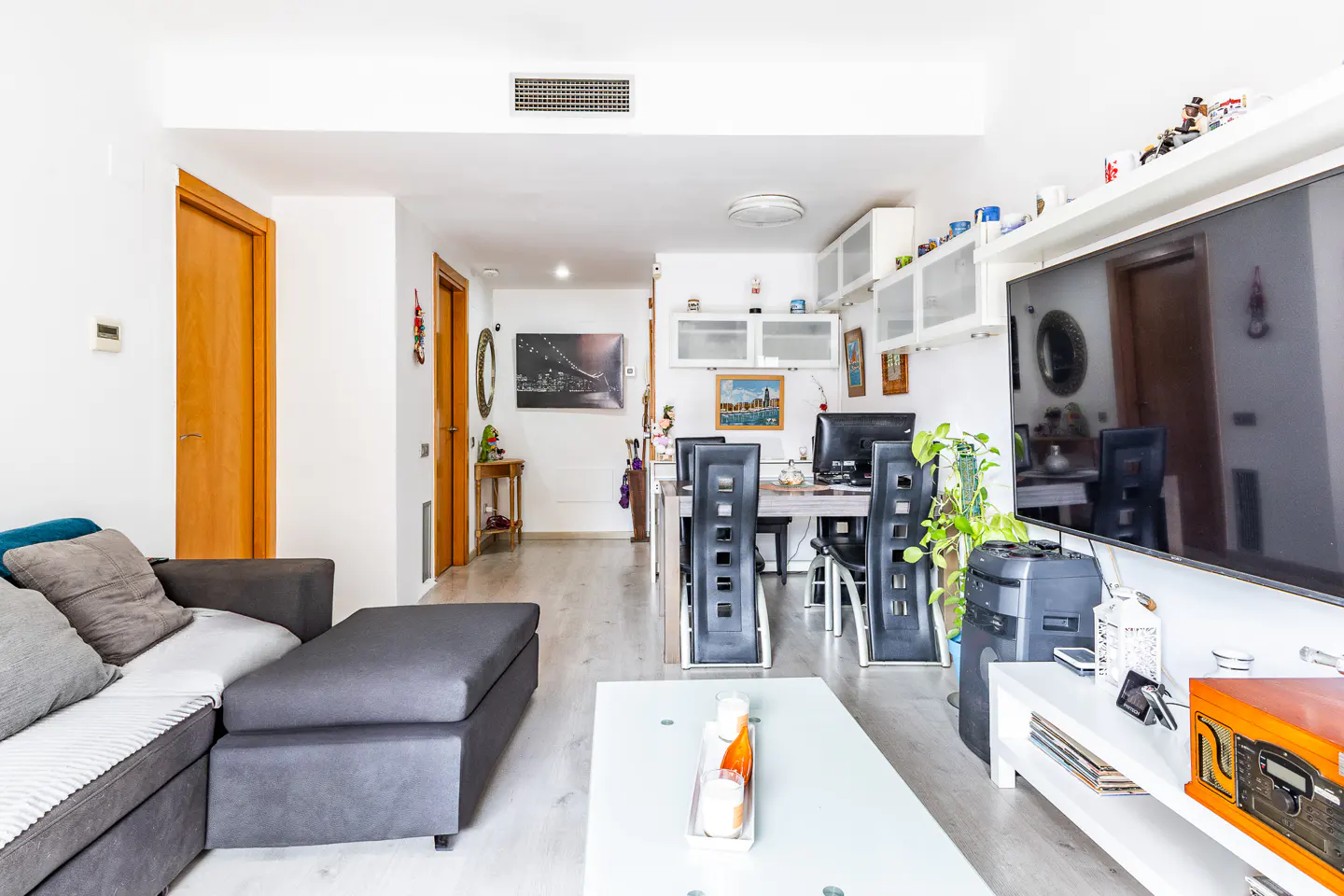 Bright, modern living room with a gray sectional sofa, white coffee table, dining set, and large TV. White walls and light wood floors.
