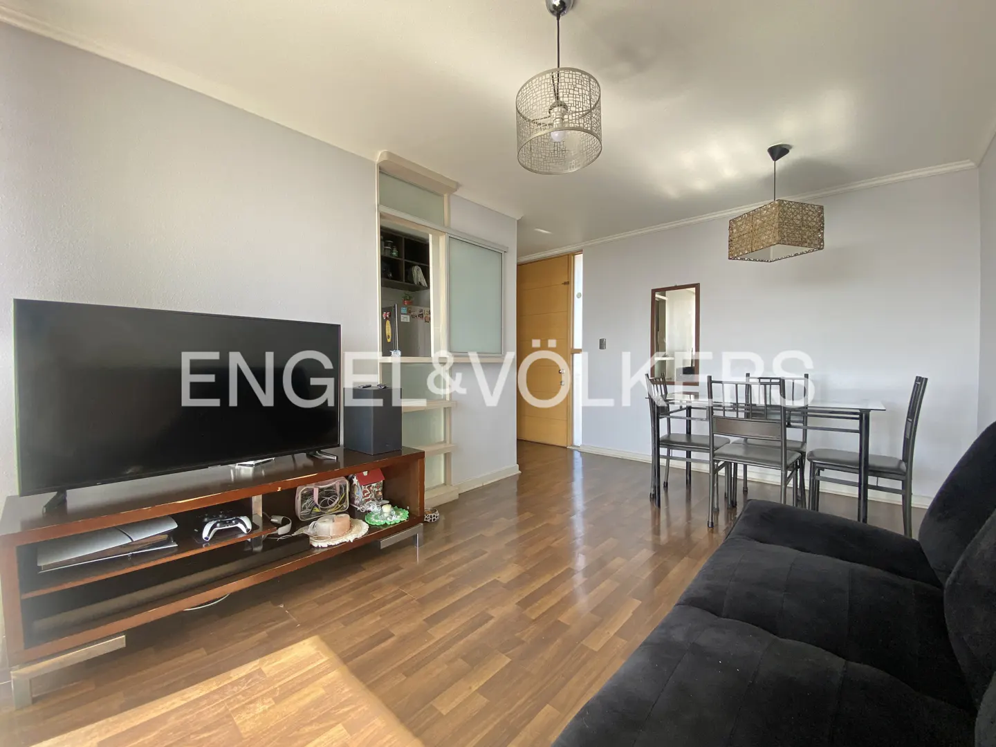 Living room with a black sofa, TV on a wooden stand, dining table with chairs, and wooden floors.