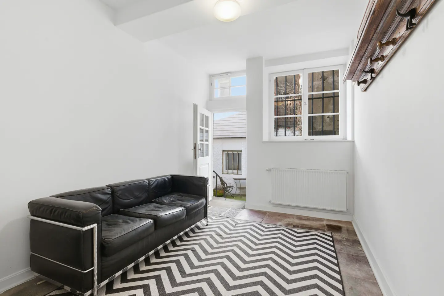 Bright room with a black leather sofa, chevron rug, and open door to a courtyard. A window with bars and a wooden coat rack are on the right.