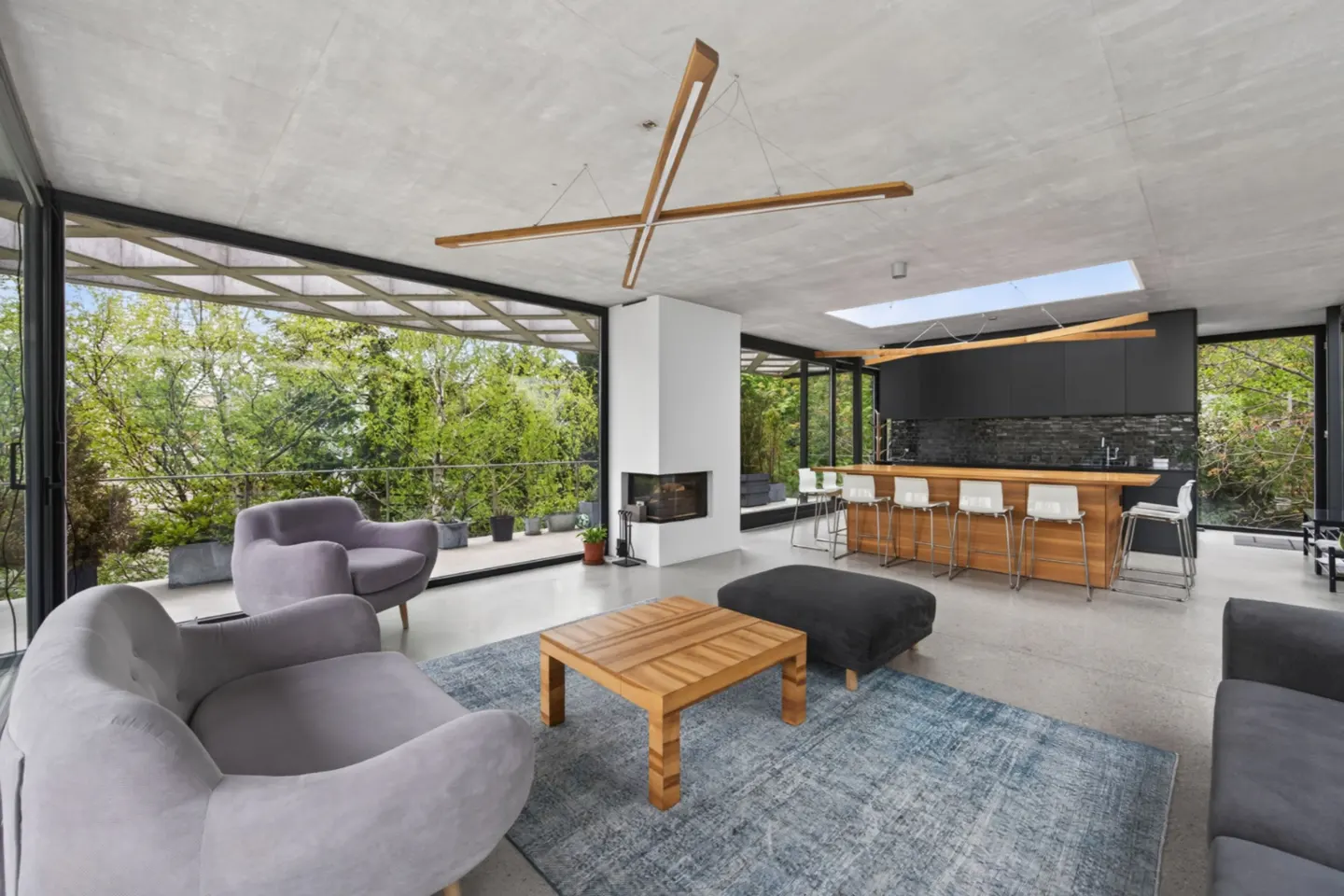 Open-concept living room with floor-to-ceiling windows, grey armchairs, wood coffee table, and kitchen bar with white stools.