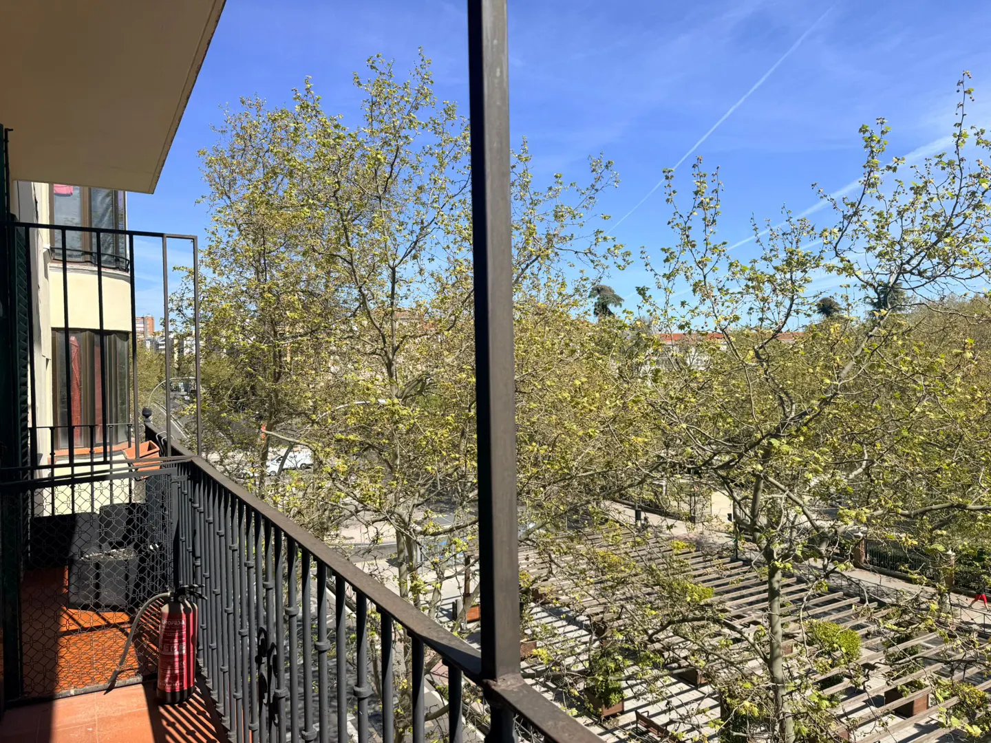 Balcony view with black iron railings overlooking trees and a blue sky. A red and white fire extinguisher is visible on the balcony.
