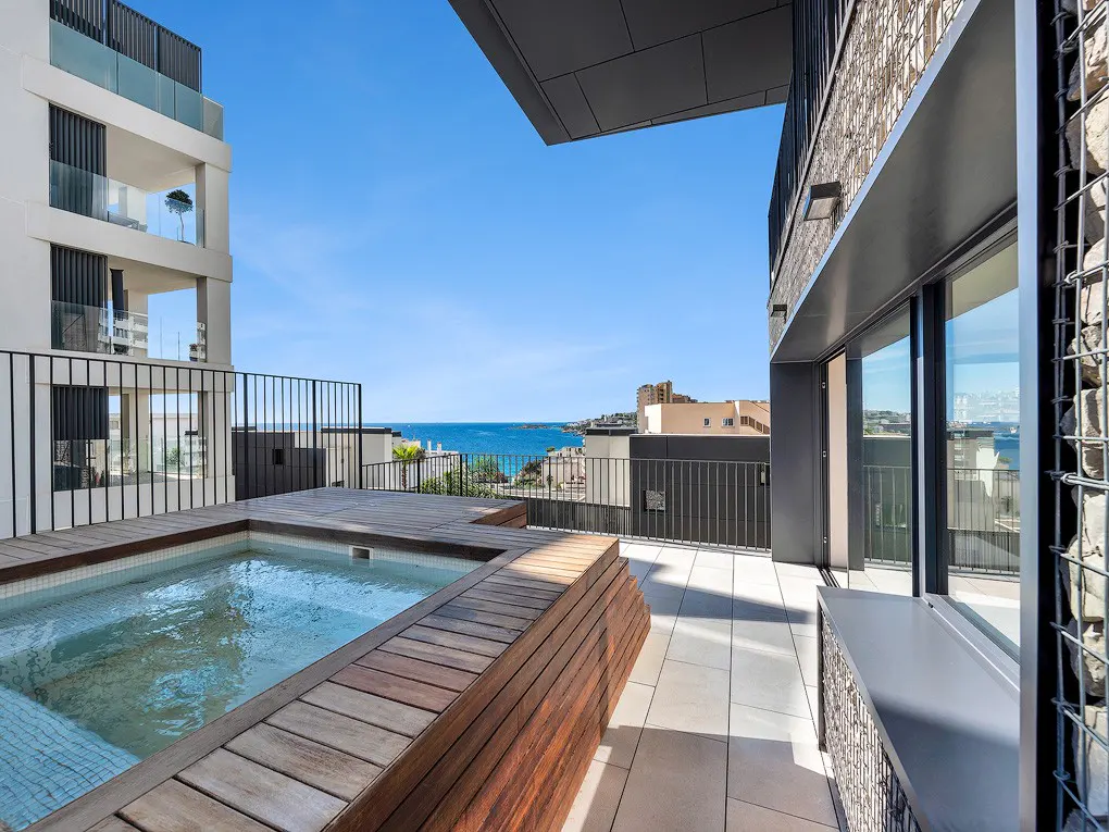 Rooftop patio with a small pool, wooden deck, and ocean view. Modern building with black railings and a clear blue sky in the background.