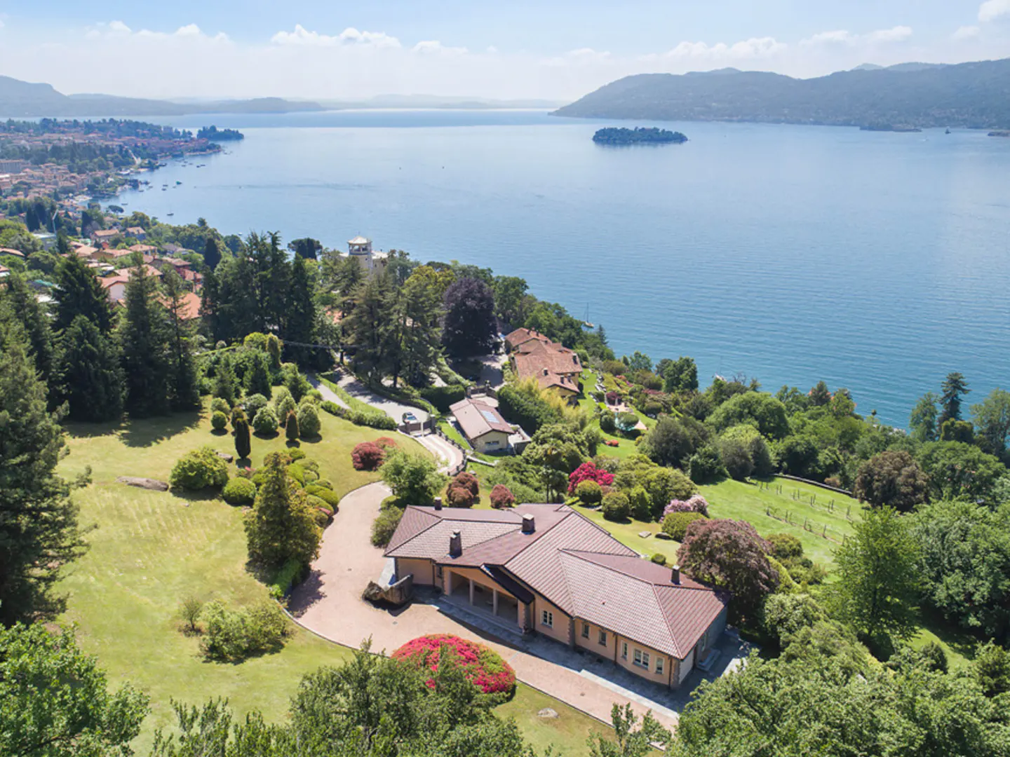 Aerial view of a tan house with a brown roof, surrounded by green trees and a blue lake with an island in the background.