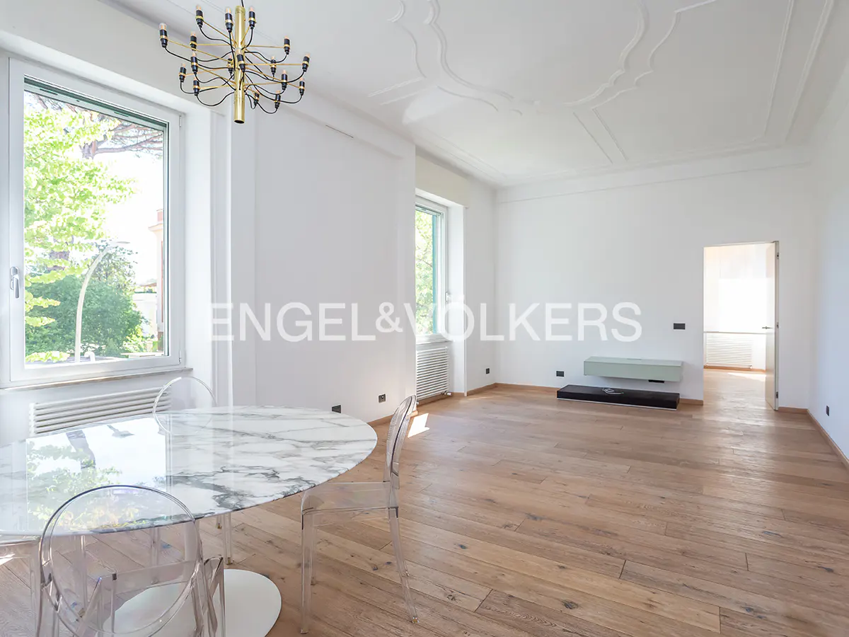 Bright, empty room with white walls, wood floors, and a marble table with clear chairs. A gold chandelier hangs from the ornate ceiling. A window shows green trees.