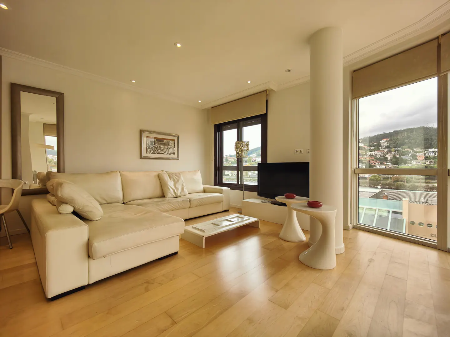 Bright living room with a cream sectional sofa, wood floors, and large windows with a view of a hillside town. A TV and white tables are also visible.