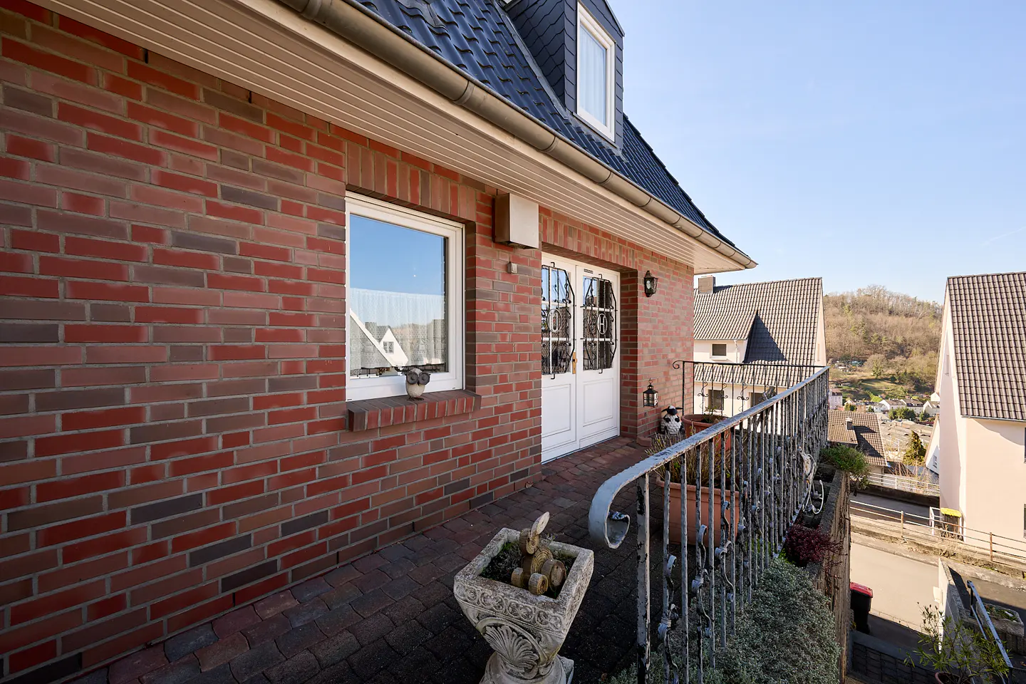 Exterior view of a red brick house with a balcony, white door, and a window. A stone planter sits on the brick patio.