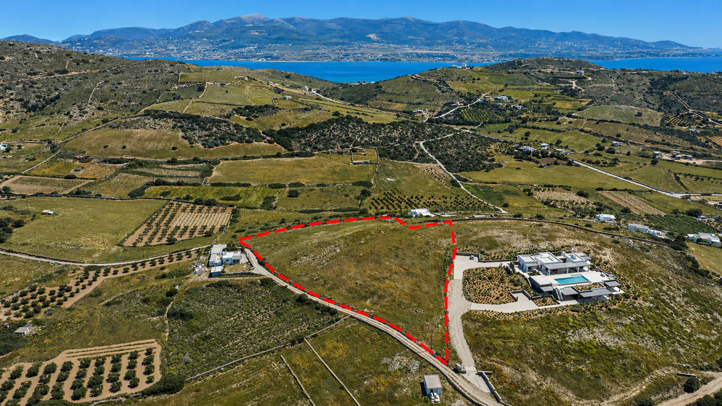 Aerial view of a vacant lot outlined in red, with a modern house nearby, set against a backdrop of green hills and a blue sea.