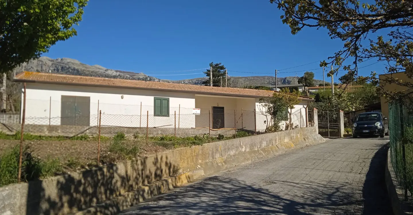 Exterior view of a one-story white house with a brown roof, green shutters, and a chain-link fence. A paved road leads to the house, with a car parked nearby. Mountains are visible in the background under a blue sky.