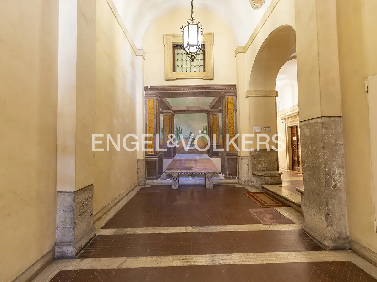 Hallway with arched ceilings, yellow walls, and a red tile floor. A stone table sits in front of a painted mural. A hanging lantern provides light.