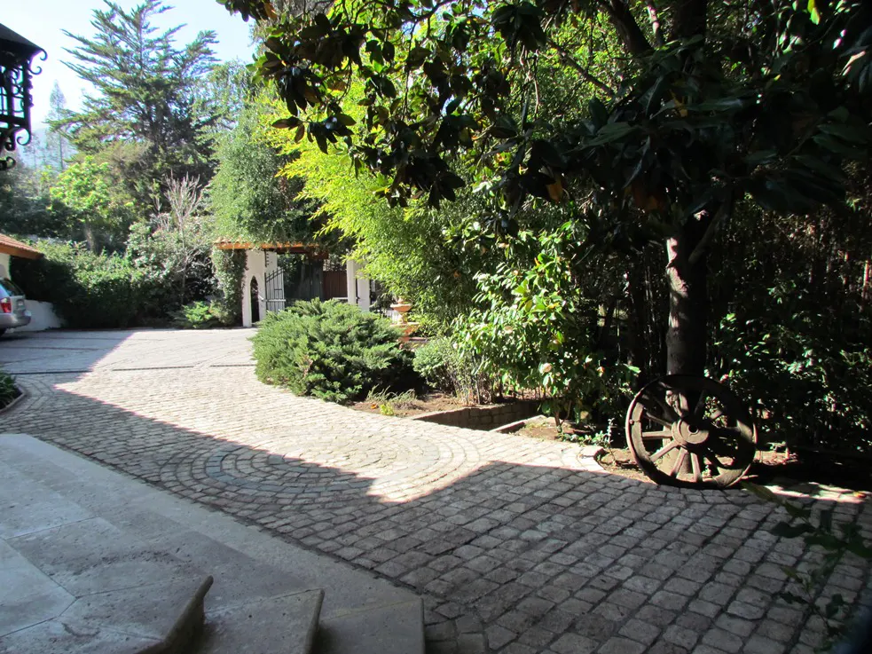 Stone driveway leading to a white gate and lush greenery. A rustic wooden wheel leans against a tree.