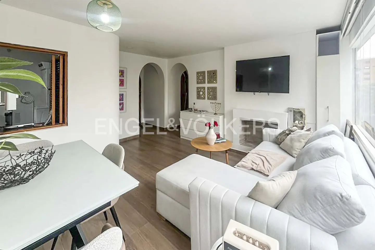 Bright living room with white walls, wood floors, and a white sofa. A dining table is on the left, and a TV is mounted above a fireplace on the right.