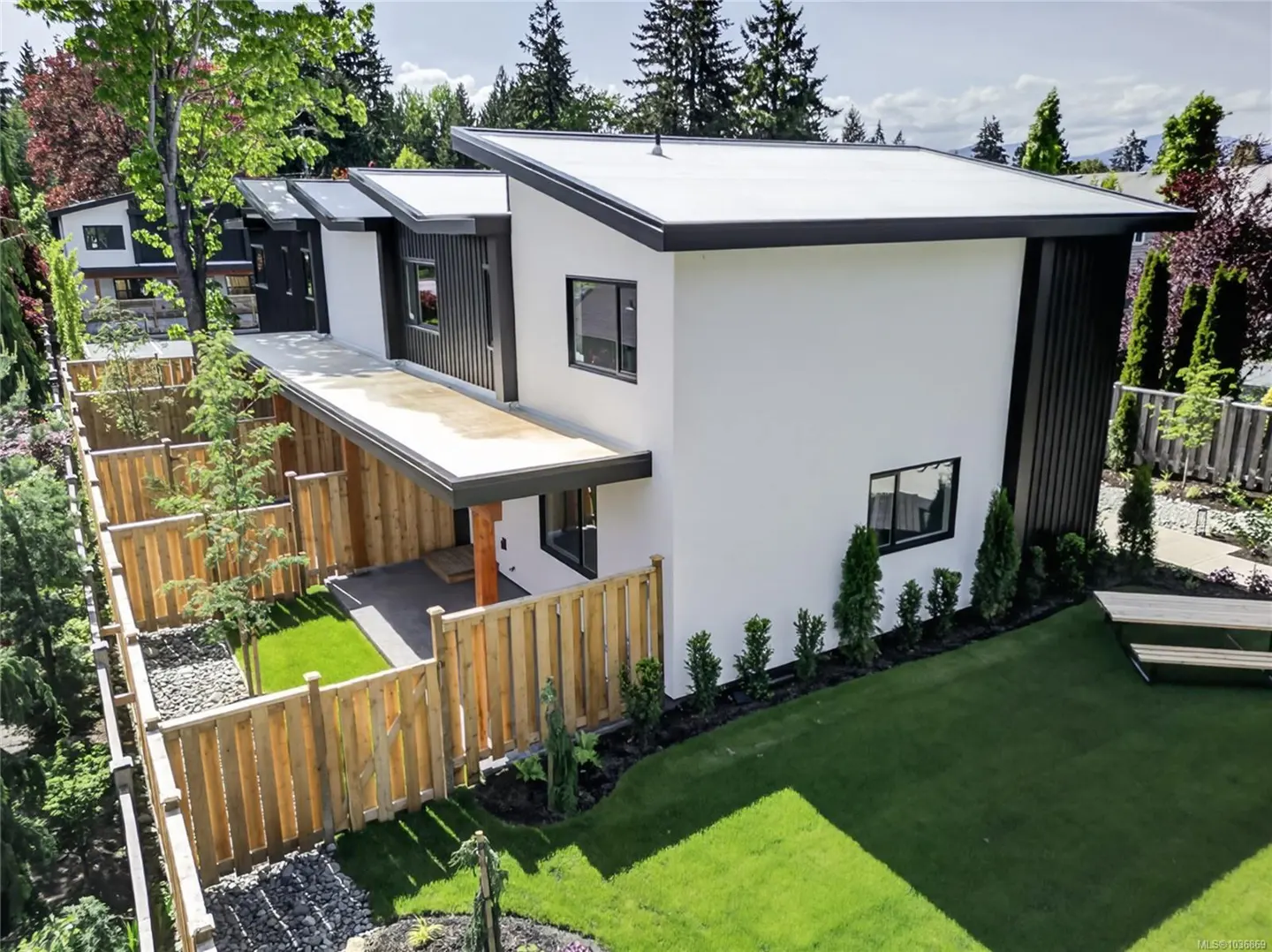 Modern white house with black trim and a flat roof, surrounded by a wooden fence and green lawn.