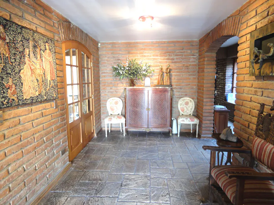Brick-walled foyer with tapestry, arched doorway, and furniture. A cabinet sits between two floral chairs.