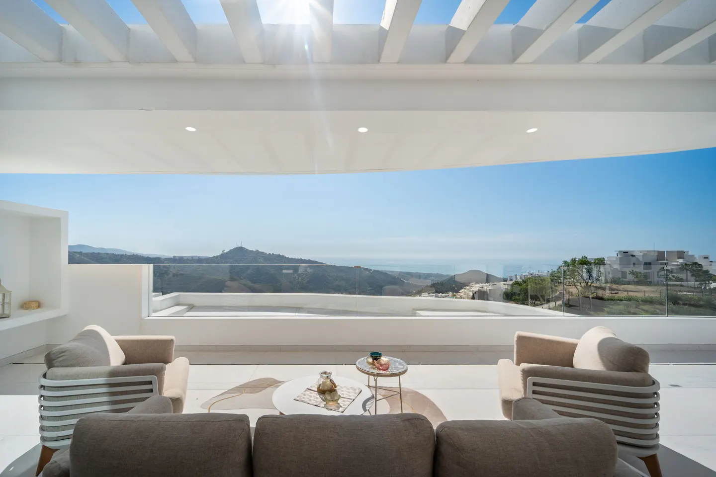 Bright, modern patio with white pergola, tan chairs, and a gray sofa. A glass railing offers views of distant mountains and a clear blue sky.