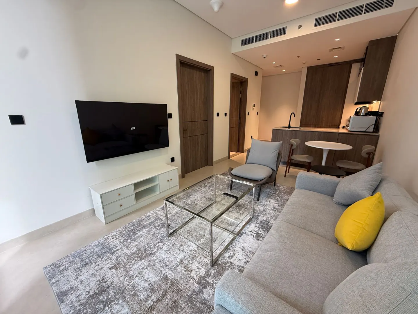 A modern apartment living room with a gray sofa, yellow pillow, glass table, TV, and kitchenette.