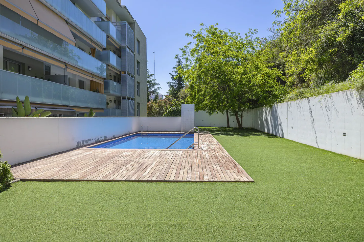 A backyard with a blue pool, wooden deck, and green grass. A white wall and building are in the background.