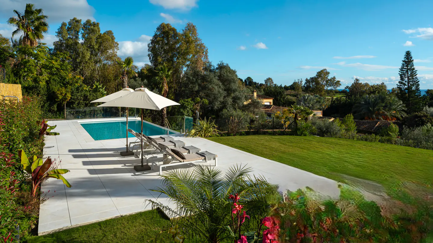 Outdoor pool area with lounge chairs, umbrellas, and green lawn overlooking trees and houses under a blue sky.