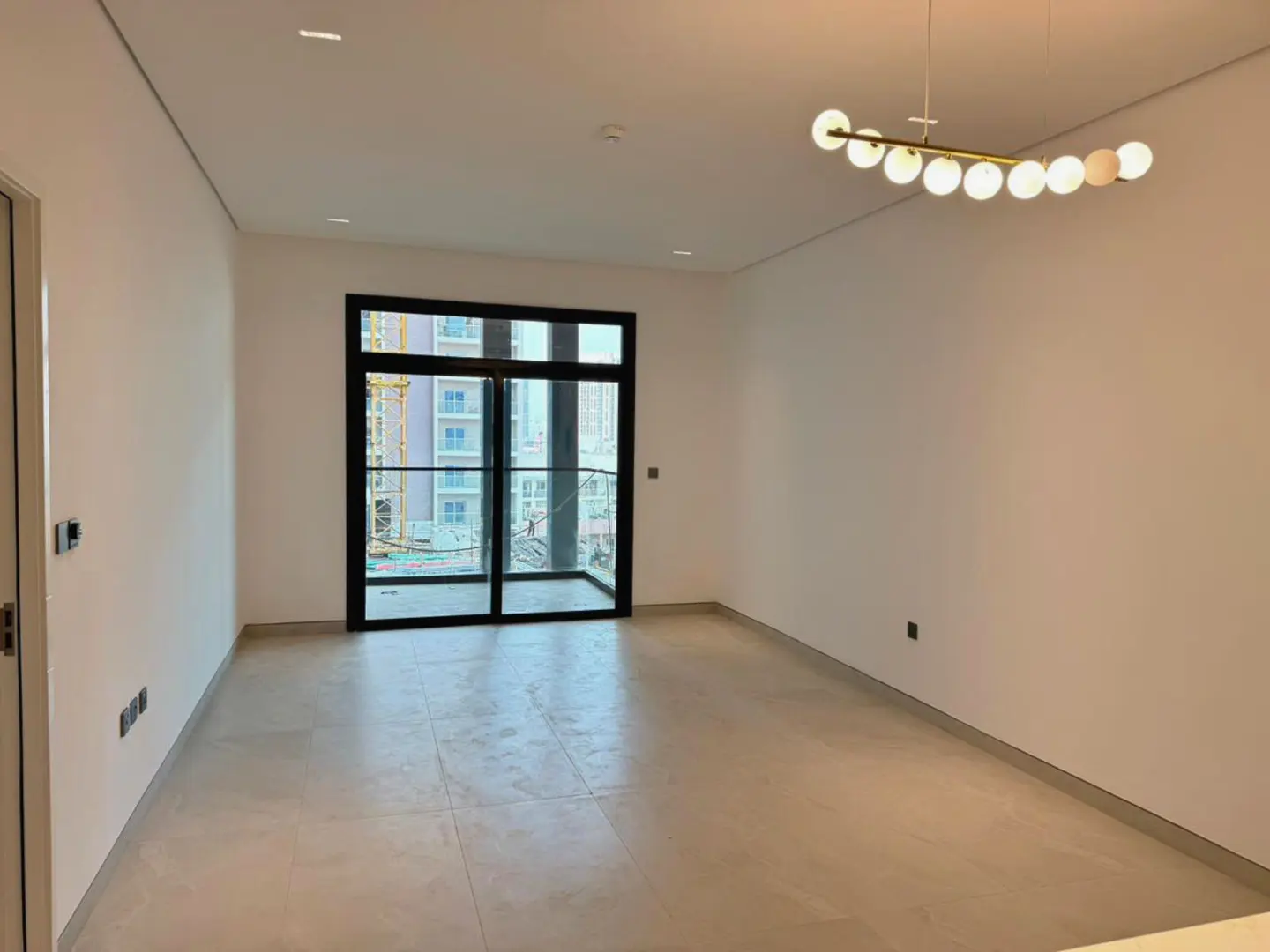 Empty room with white walls, light tile floor, and black-framed sliding glass doors to a balcony. A modern light fixture hangs from the ceiling.