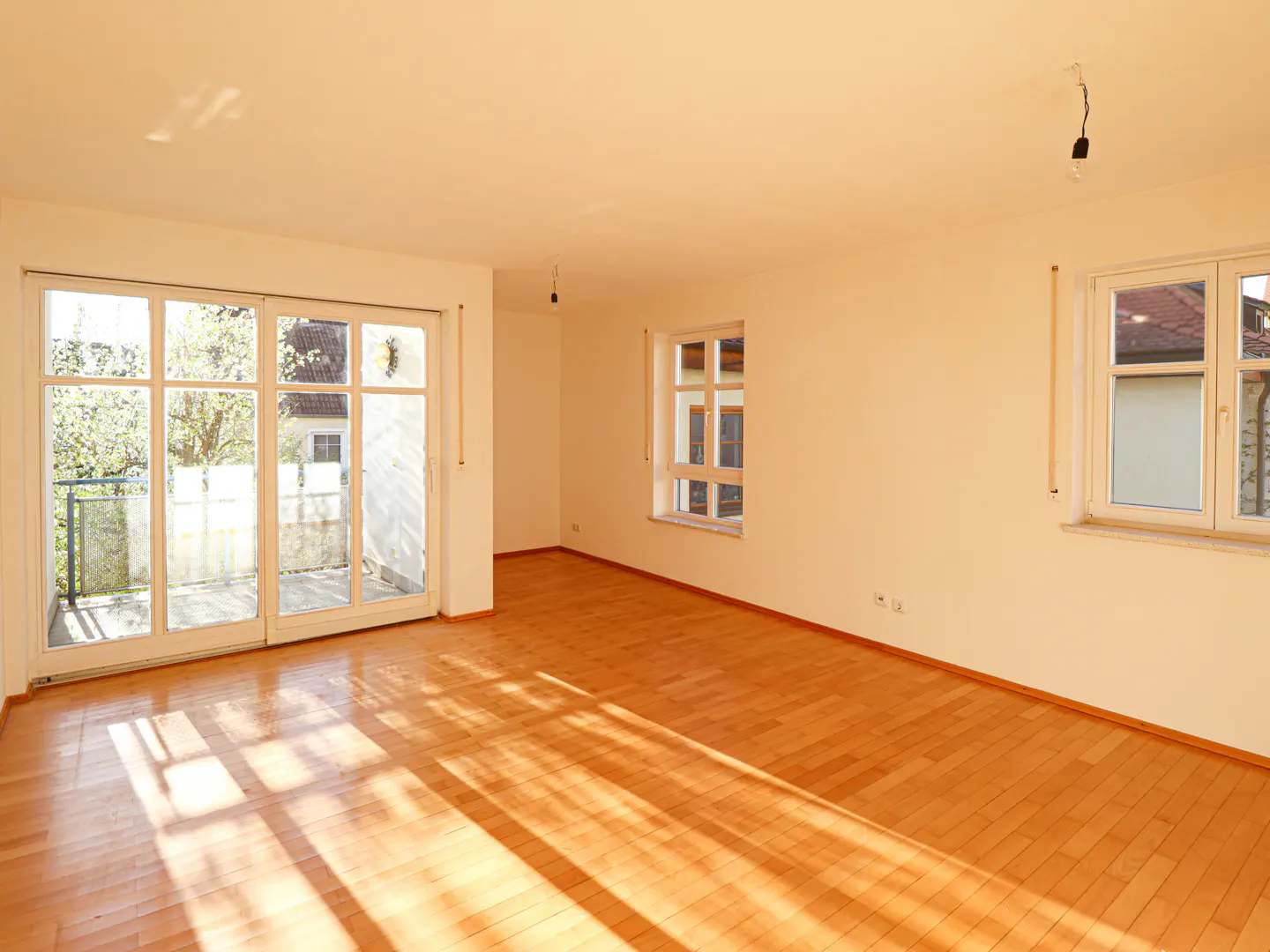 Empty room with hardwood floors, white walls, and natural light. Sliding glass doors lead to a balcony. Two windows offer outside views.