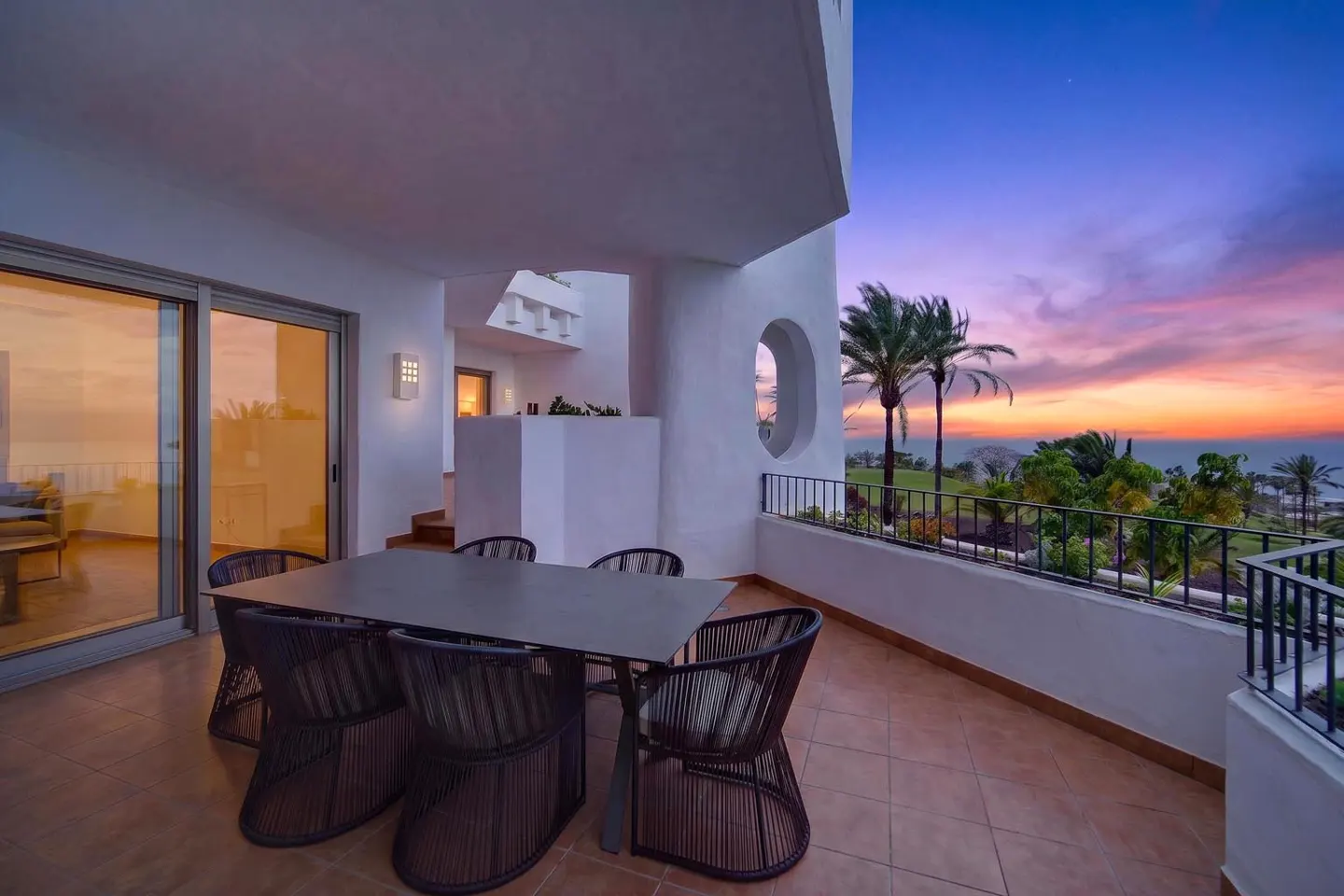 Outdoor patio with a table and chairs overlooking a sunset view with palm trees and ocean.