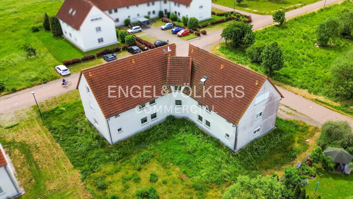 Aerial view of an L-shaped white apartment building with a brown tiled roof, surrounded by green lawns and a parking lot.