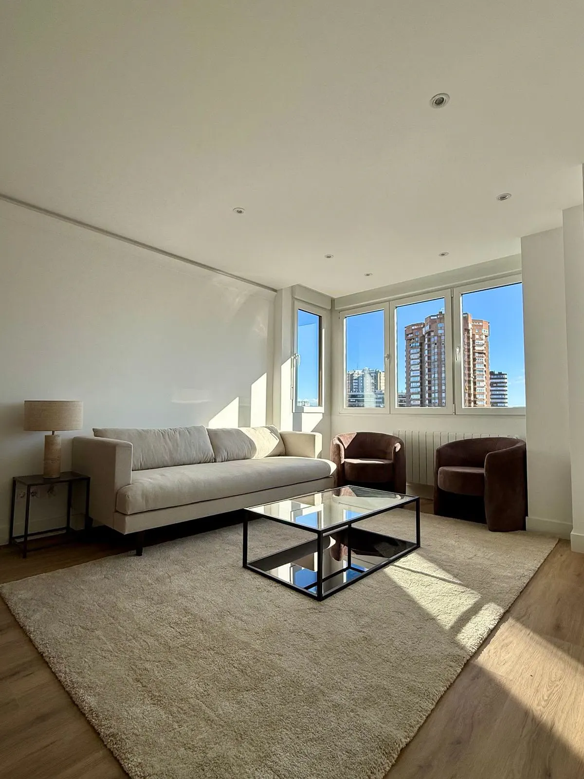 Bright living room with a long beige sofa, glass coffee table, two brown armchairs, and city views through large windows.