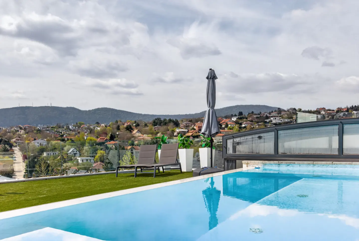 Rooftop pool with blue water, two lounge chairs, and a closed gray umbrella. A city and mountains are in the background.