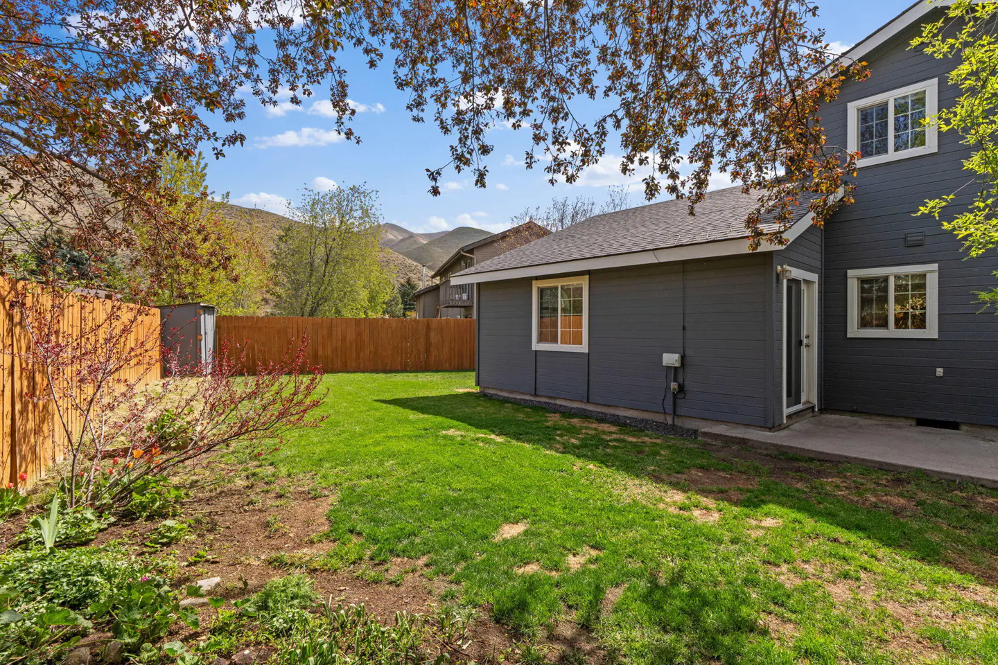 A backyard with green grass, a wooden fence, and a gray house with white trim. Mountains are visible in the background.